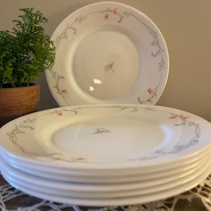 May include: A stack of six white ceramic plates with a delicate floral design in shades of pink and gray. The plates have a simple, elegant design and would be perfect for a casual dinner party.