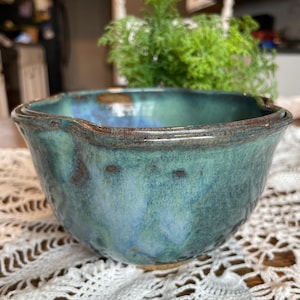 May include: A handmade ceramic bowl with a turquoise and blue glaze. The bowl has a unique shape with a pouring spout and a dark brown rim. The interior shows a blend of blue and green hues, creating a textured appearance. The bowl sits on a white lace tablecloth.