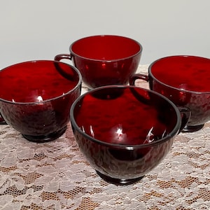 May include: Four ruby red glass teacups with handles. The cups have a textured surface and a deep, rich color. The cups are arranged on a lace tablecloth. The cups are likely vintage or antique.