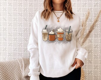 May include: White sweatshirt with a graphic of four different coffee drinks with whipped cream on top. The drinks are a latte, a pumpkin spice latte, a caramel macchiato, and an iced coffee.
