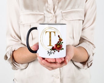 May include: White ceramic mug with a black rim and handle. The mug features a gold circle with the letter "T" in gold and a floral design with red and gold roses. The name "Tyler" is written in black below the circle.