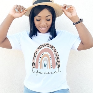 Op de afbeelding: Wit T-shirt met een regenbooggrafiek met een luipaardprint. De regenboog heeft een kleurenschema van roze, oranje, geel en blauw. De tekst "life coach" is onder de regenboog gedrukt.