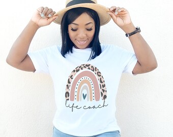 May include: White t-shirt with a rainbow graphic featuring a leopard print design. The rainbow has a pink, orange, yellow, and blue color scheme. The text "life coach" is printed below the rainbow.