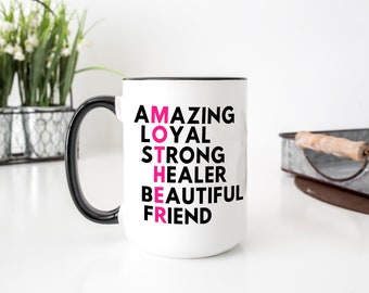 May include: White ceramic coffee mug with a black rim and handle. The mug has the word "MOTHER" spelled vertically in pink letters, with the words "AMAZING", "LOYAL", "STRONG", "HEALER", "BEAUTIFUL", and "FRIEND" spelled out vertically around it.