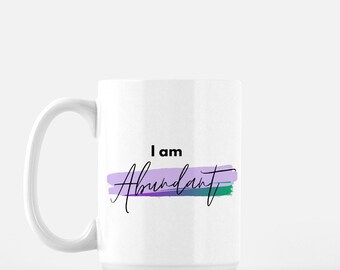 May include: White ceramic coffee mug with a black handle. The mug has a purple, blue, and green watercolor design with the text "I am Abundant" in black.