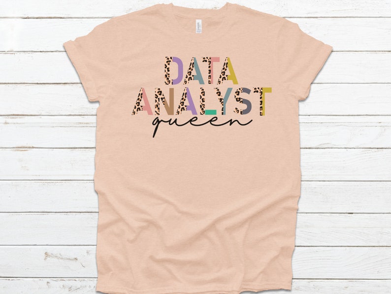 May include: A peach-colored t-shirt with the text "DATA ANALYST queen" in colorful letters with a leopard print pattern.