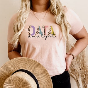 以下が含まれることがあります： レオパード柄のカラフルな文字で「DATA analyst」と書かれたピンクのTシャツです。