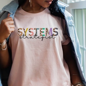 Op de afbeelding: Een roze T-shirt met de tekst "SYSTEMS strategist" in kleurrijke letters met een luipaardprint.