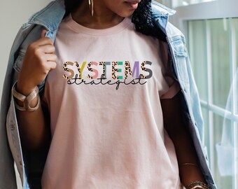 May include: A pink t-shirt with the text "SYSTEMS strategist" in colorful letters with a leopard print pattern.
