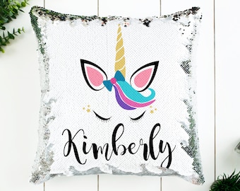 May include: A white sequin pillow with a unicorn face and the name Kimberly printed on it. The unicorn has a gold horn, pink ears, a blue and purple mane, and closed eyes.