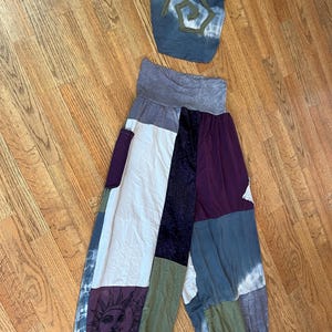 May include: A two-piece outfit consisting of a halter top and loose-fitting pants. The top is blue with a tie-dye effect and a spiral patch. The pants feature a patchwork design in white, purple, blue, and olive green, including a sun graphic.