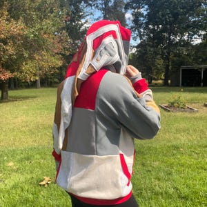 May include: A long-sleeve hooded sweatshirt featuring a patchwork design of red, gray, tan, and white fabric panels. The garment includes a large hood with layered fabric detailing, long sleeves with ribbed cuffs, and a red hemline at the bottom.