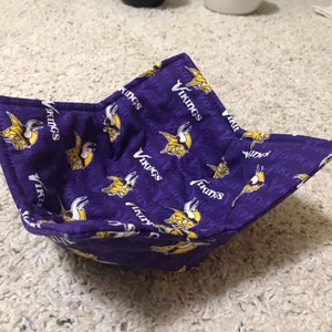 Puede incluir: Tazón de tela morado con un patrón repetido de cascos vikingos amarillos y la palabra "Vikingos" sobre un fondo morado.