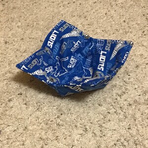 Pode incluir: Uma tigela de tecido azul com uma estampa branca da palavra "Lions" repetida por toda parte. A tigela é dobrada e tem uma ligeira curva.