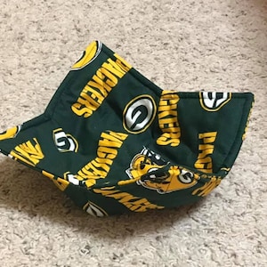Op de afbeelding: Een groene en gele stoffen kom-cozy met de woorden "PACKERS" en het logo van het team. De cozy is ontworpen om een kom vast te houden en het eten warm te houden. De stof heeft een herhalend patroon van de naam en het logo van het team.