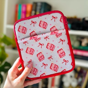 Puede incluir: Una funda cuadrada y acolchada para tablet con cremallera y ribete rojos. La tela gris claro está estampada con ilustraciones de libros rojos, lazos y pequeños corazones rosas. El texto en las ilustraciones de libros dice "Hot Girls Read".