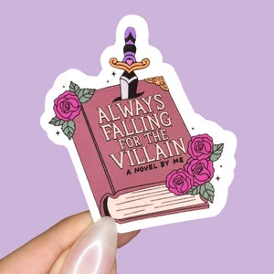 Puede incluir: Una pegatina de libro rosa con una daga que sobresale de la parte superior. La portada del libro dice "Always Falling for the Villain, A Novel by Me" y tiene rosas rosas en el lateral.