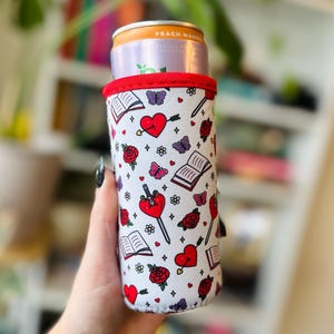 May include: A white beverage can cooler with a red trim, featuring a repeating pattern of red hearts pierced by swords, roses, butterflies, and open books. The cooler holds a can of Peach Mango flavored beverage.
