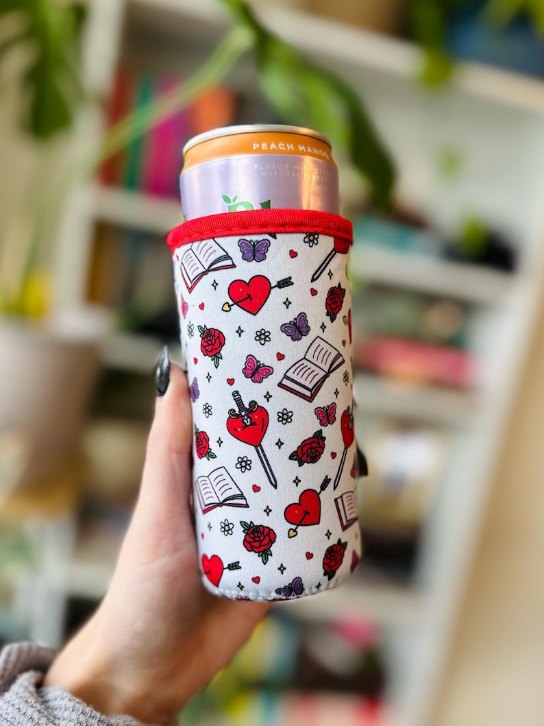 May include: A white beverage can cooler with a red trim, featuring a repeating pattern of red hearts pierced by swords, roses, butterflies, and open books. The cooler holds a can of Peach Mango flavored beverage.
