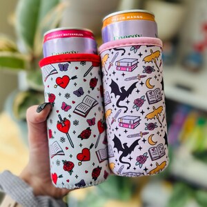 May include: Two beverage can coolers with fantasy-themed patterns. One features hearts, arrows, roses, and butterflies. The other has dragons, swords, books, and moons. Both have a white background with red and pink accents.