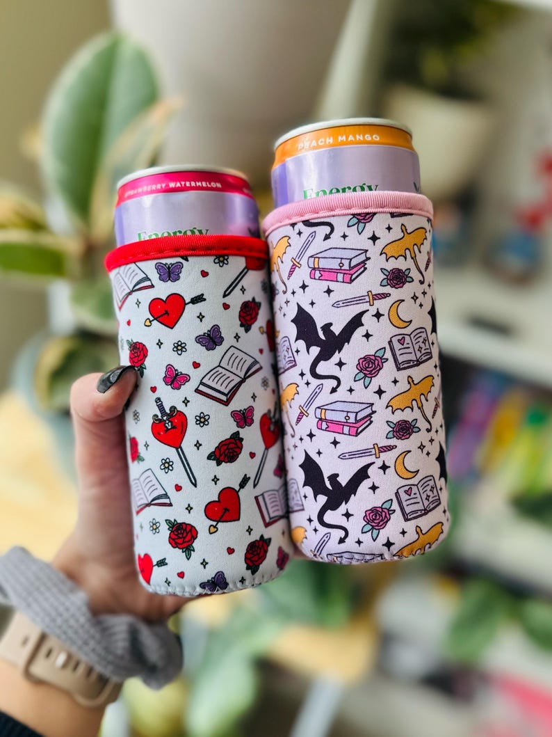 May include: Two beverage can coolers with fantasy-themed patterns. One features hearts, arrows, roses, and butterflies. The other has dragons, swords, books, and moons. Both have a white background with red and pink accents.