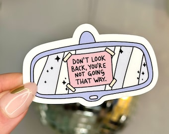 May include: A white sticker featuring a cartoon illustration of a rearview mirror. The mirror reflects a pink note that reads "Don't look back, you're not going that way."