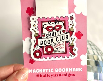 Könnte beinhalten: Ein rosa magnetischer Lesezeichen mit einem weißen Aufkleber, der eine rote und weiße Illustration einer Maske, Handschellen, Chilischoten und den Text "Smutty Book Club - Admit One - Good Girls Only" zeigt. Das Lesezeichen trägt außerdem den Text "Magnetic Bookmark @haileylizdesigns".