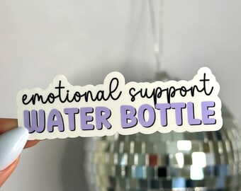 Peut inclure: Un autocollant blanc avec le texte "emotional support water bottle" en lettres noires et violettes. L'autocollant est tenu par une main.