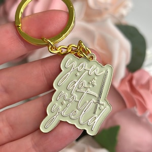 Puede incluir: Un llavero de metal verde claro con una cadena dorada y un diseño de espada. El llavero tiene las palabras "you do not yield" escritas en blanco.