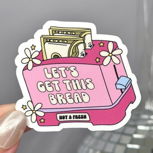 Può includere: Un tostapane rosa con un mucchio di soldi dentro, circondato da fiori bianchi e stelle. Il tostapane ha la scritta "Let's Get This Bread" e "Hot & Fresh" stampata su di esso.