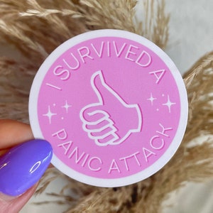 Puede incluir: Una pegatina redonda rosa con un contorno blanco. La pegatina tiene un icono de pulgar hacia arriba blanco y el texto "Sobreviví a un ataque de pánico" en blanco.