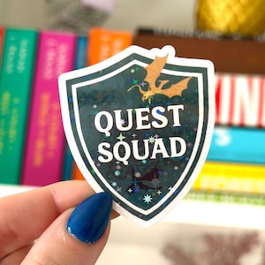 May include: A green and gold shield sticker with a dragon and the text "QUEST SQUAD" in white letters. The shield has a glittery finish.