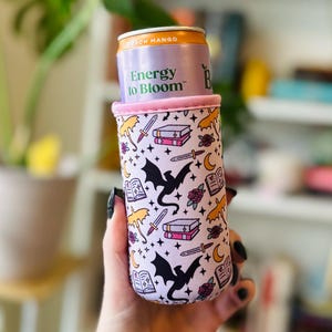 May include: A white and pink can cooler with a fantasy theme. The cooler features black dragons, books, swords, roses, and umbrellas. The can inside reads "Energy to Bloom" and "Peach Mango".