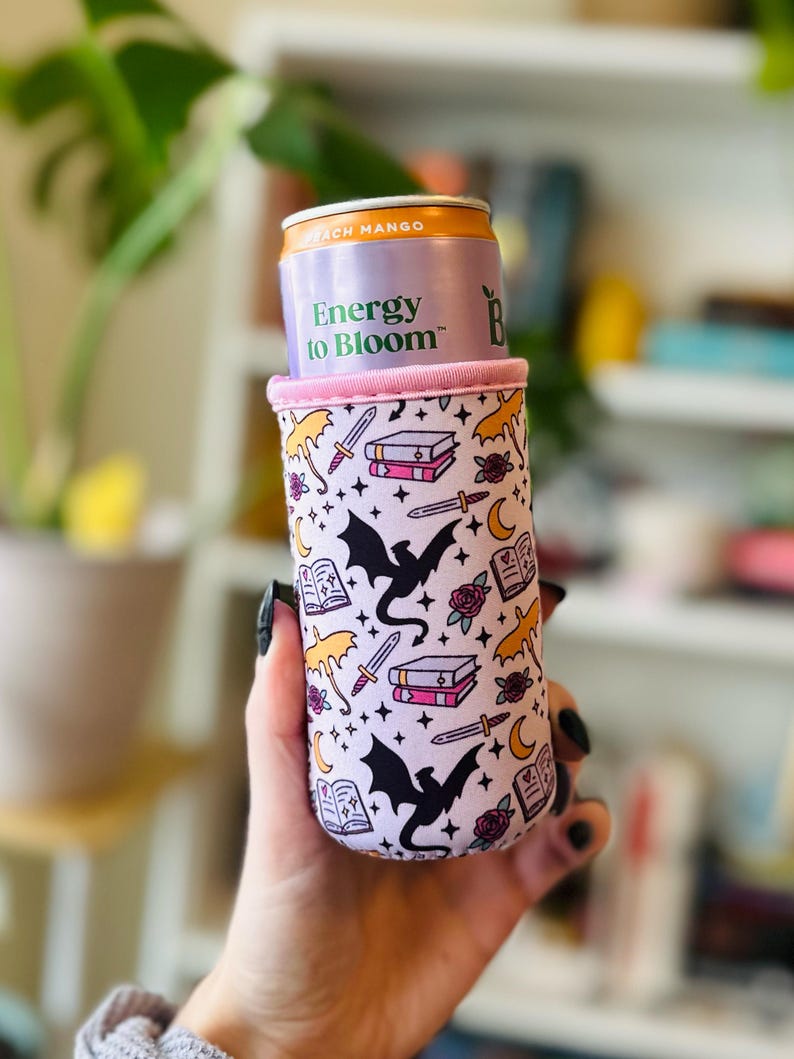 May include: A white and pink can cooler with a fantasy theme. The cooler features black dragons, books, swords, roses, and umbrellas. The can inside reads "Energy to Bloom" and "Peach Mango".