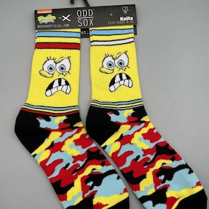 May include: Pair of yellow Odd Sox featuring a cartoon character's face with a surprised expression. The socks have a black heel and toe, and a colorful camouflage pattern in red, yellow, and blue. The socks fit sizes 6-13.