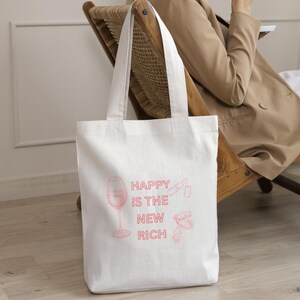Könnte beinhalten: Weißer Canvas-Tote-Bag mit rosa Grafiktext und Illustrationen. Der Text lautet "HAPPY IS THE NEW RICH" mit Bildern eines Weinglases, Lippenstifts und Austern. Die Tasche hat zwei weiße Griffe und wird von einer Person gehalten.