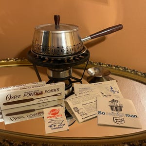 May include: A vintage fondue set with a stainless steel pot, wooden handle, and burner. Includes boxes of fondue forks and printed recipe cards. The set is displayed on a mirrored surface.