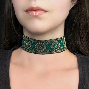 May include: A black choker necklace with a geometric pattern in green and gold. The choker is worn around the neck and features a detailed woven design. The necklace is a stylish accessory.
