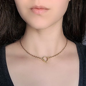 以下が含まれることがあります： トグルクラスプ付きのゴールドカラーのネックレス。 ネックレスはチョーカータイプで、首の近くにあります。 チェーンは小さなリンクされたセグメントでできています。 クラスプは、バークロージャー付きの円形リングです。