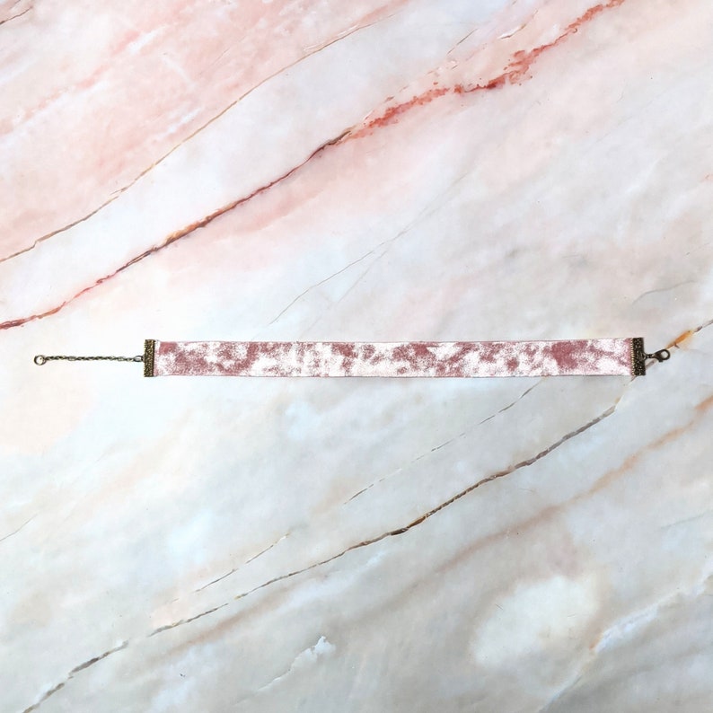 Op de afbeelding: Een roze fluwelen choker met een zilveren ketting en metalen sluitingen.