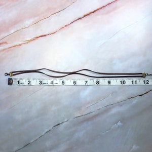 May include: A brown leather cord necklace with gold-toned clasps. The necklace is measured on a ruler and is approximately 11 inches long.
