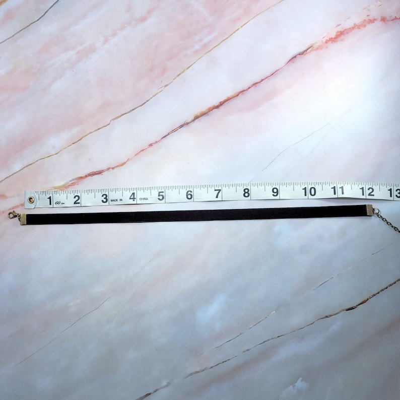 Puede incluir: Un collar gargantilla negro, de aproximadamente 30 cm de largo, con un cierre y una cadena de color plateado. El collar est&aacute; hecho de un material fino y aterciopelado. El fondo es un patr&oacute;n de m&aacute;rmol rosa claro y blanco.