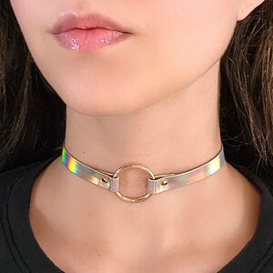 May include: A holographic choker necklace with a gold ring accent. The choker is made of a thin, flexible material and has a subtle iridescent sheen.