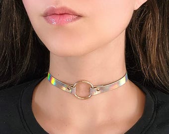 Op de afbeelding: Een holografische choker ketting met een gouden ring accent. De choker is gemaakt van een dun, flexibel materiaal en heeft een subtiele iriserende glans.