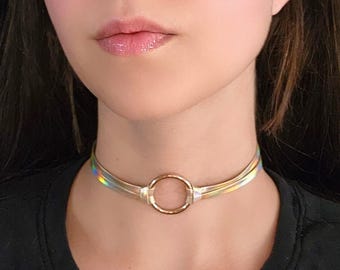 Op de afbeelding: Een iriserende choker ketting met een gouden cirkel hanger. De choker is gemaakt van een dun, flexibel materiaal en heeft een subtiele glans.