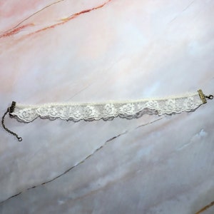 K&ouml;nnte beinhalten: Eine wei&szlig;e Spitzen-Choker-Halskette mit einem zarten Blumenmuster und einem Metallverschluss.