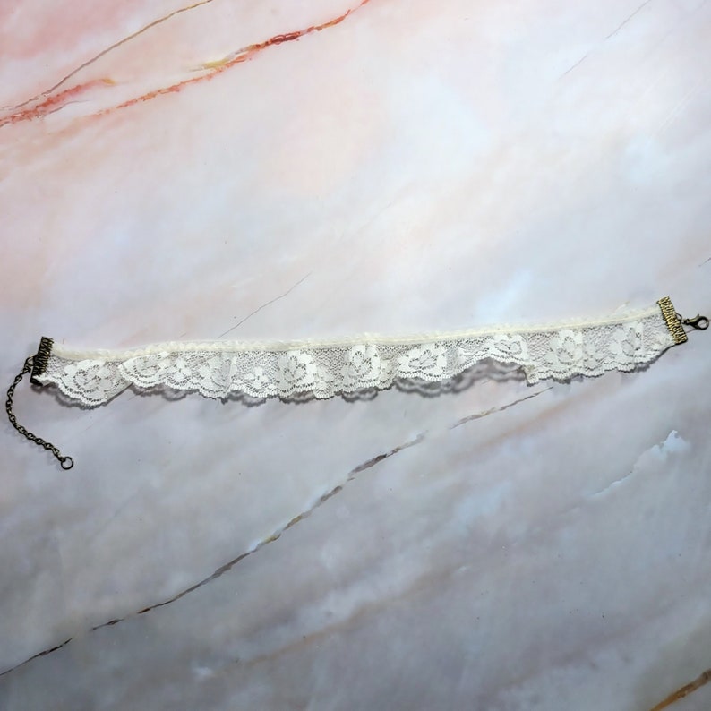 K&ouml;nnte beinhalten: Eine wei&szlig;e Spitzen-Choker-Halskette mit einem zarten Blumenmuster und einem Metallverschluss.