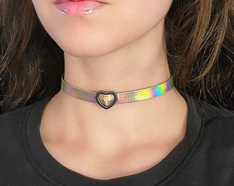 Op de afbeelding: Een holografische, iriserende choker met een zwarte hartvormige hanger. De choker is gemaakt van een dun, flexibel materiaal en heeft een eenvoudig, elegant ontwerp.