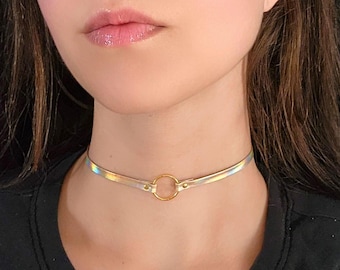 Op de afbeelding: Een choker ketting met een dunne, iriserende band en een goudkleurige ring in het midden. De band lijkt van kleur te veranderen in het licht. De ketting wordt om de nek gedragen.