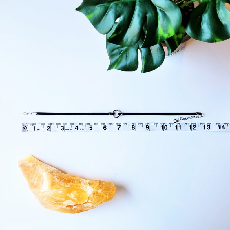 May include: A black choker necklace with a silver circle pendant. The necklace is measured on a ruler, showing it is 17.8 cm long.